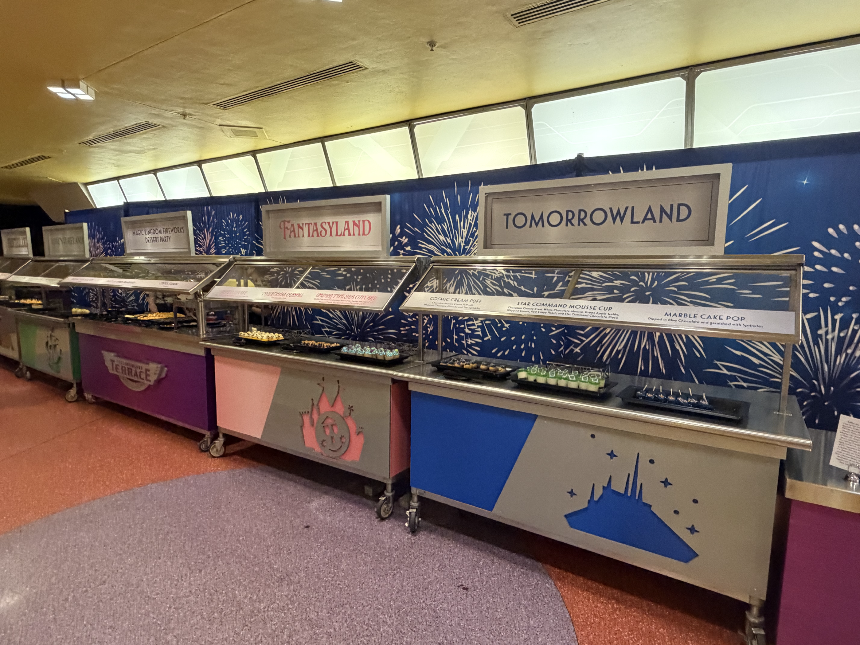 4 buffet tables standing next to each other, each with emblems from the different Magic Kingdom lands. Each table has an assortment of desserts. Behind the tables is a wallpaper of fireworks.