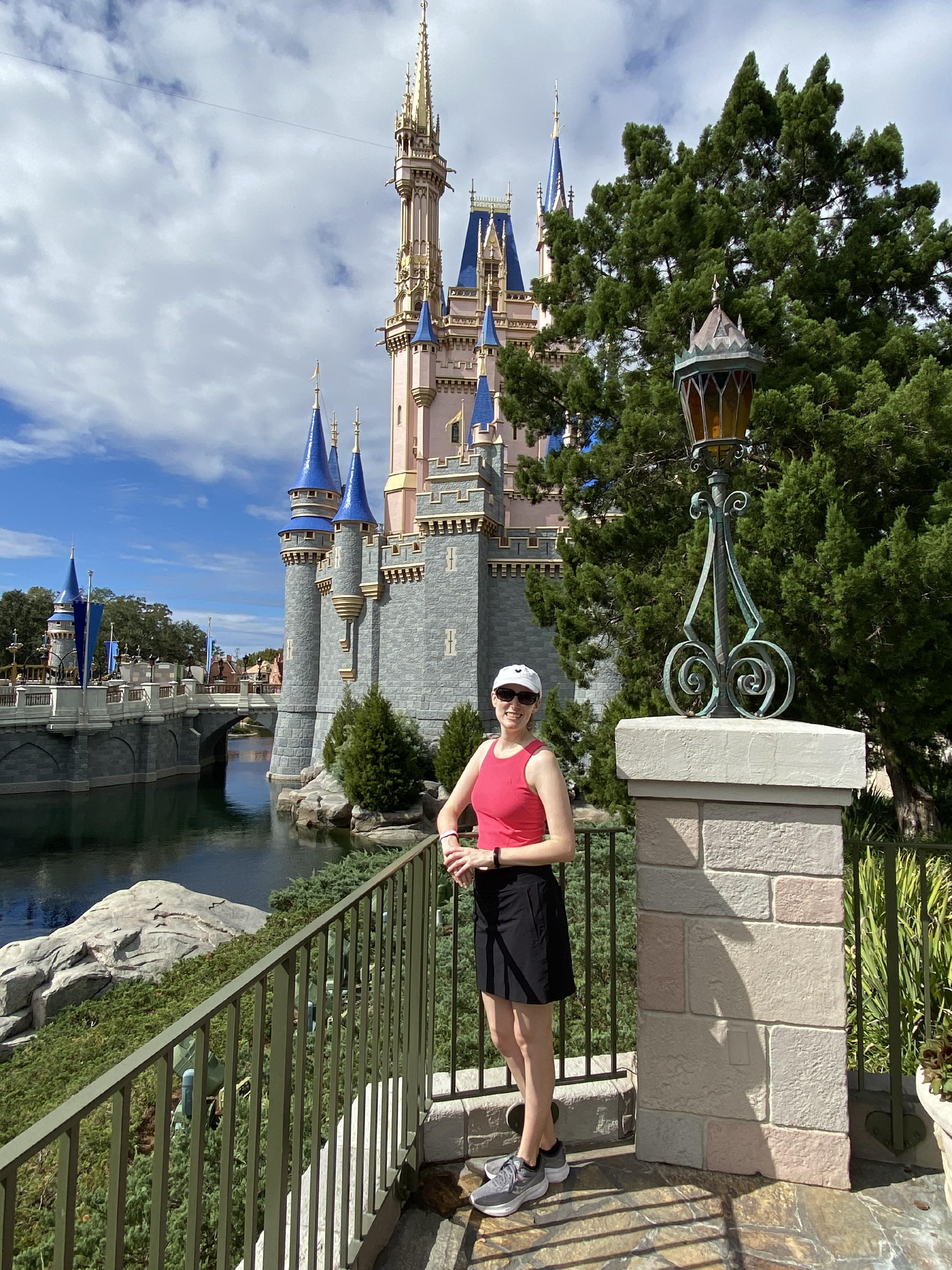 A photo of a blonde woman standing at the side of the Disney Castle. She is wearing a pink top, sunglasses, and a baseball cap.